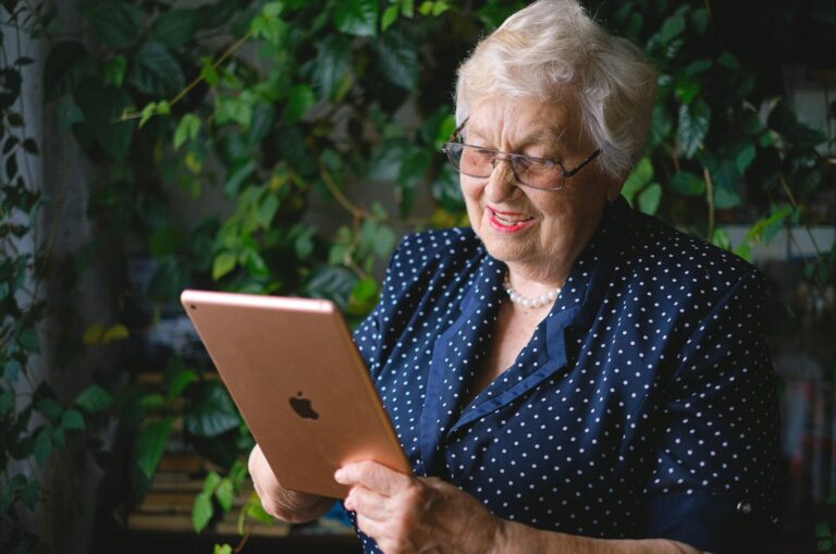 A photograph of a senior woman sat down and smiling at her tablet device.