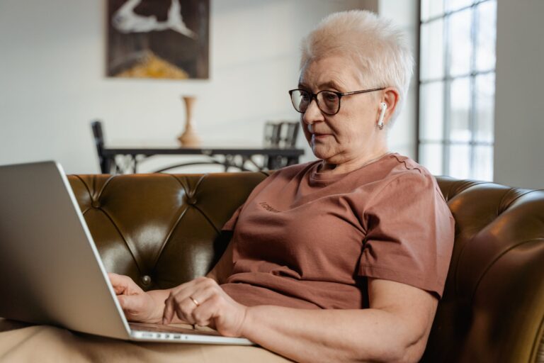 A photograph of an older woman with short hair, glasses and wireless earphones sat on a sofa looking a a laptop on her lap.
