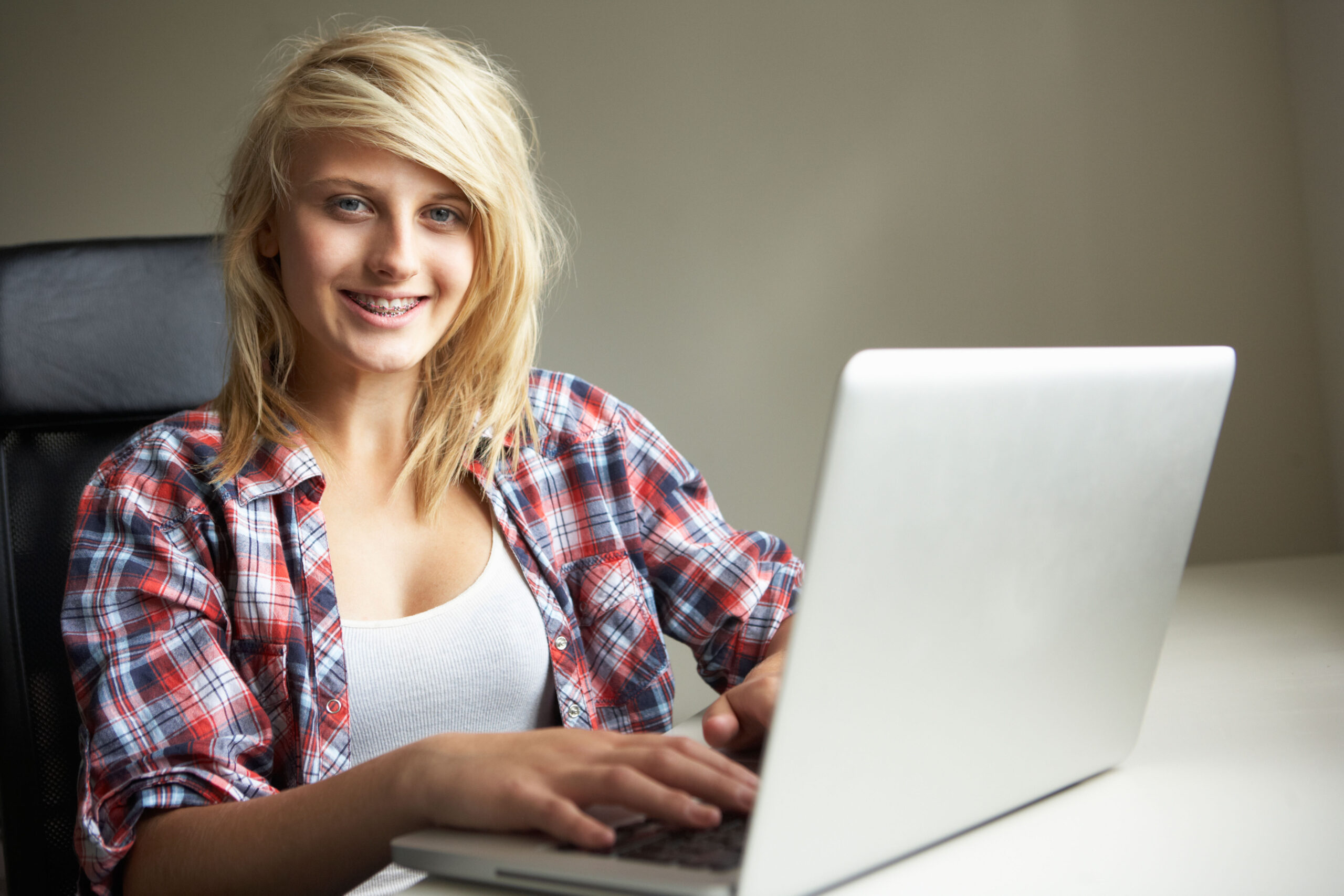 A photograph of a smiling teenage girl sat down at a desk using a laptop.