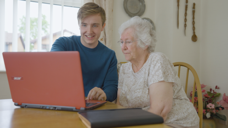 A photograph of a young man Digital Champion showing a senior lady how to use a laptop. They are both sat down at a desk in a living room.