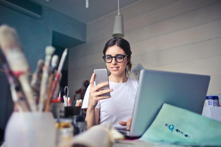 A photograph of a young woman sat behind a laptop in an art studio. She is wearing glasses looking at her phone. In the foreground there is a pot of paintbrushes.