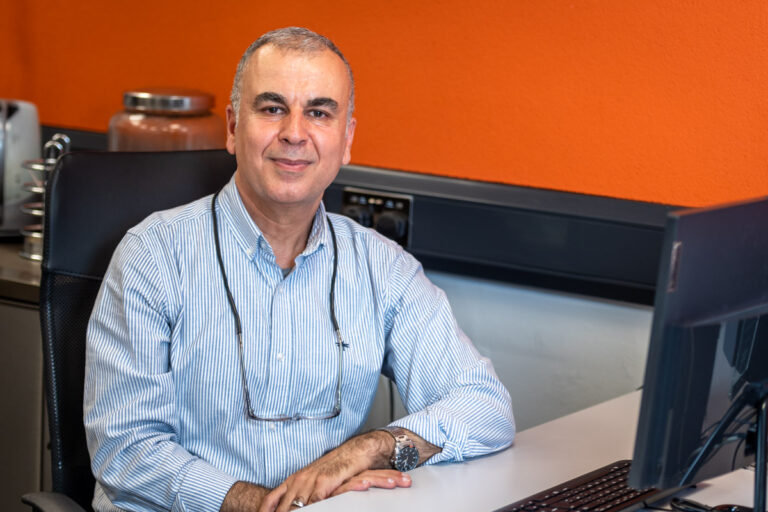 A photograph of Finance Officer, Awder. He is sat at a desk with a computer in front of him. His glasses hang around his neck suspended with a strap as he smiles at the camera while crossing his arms, rested on the desk.