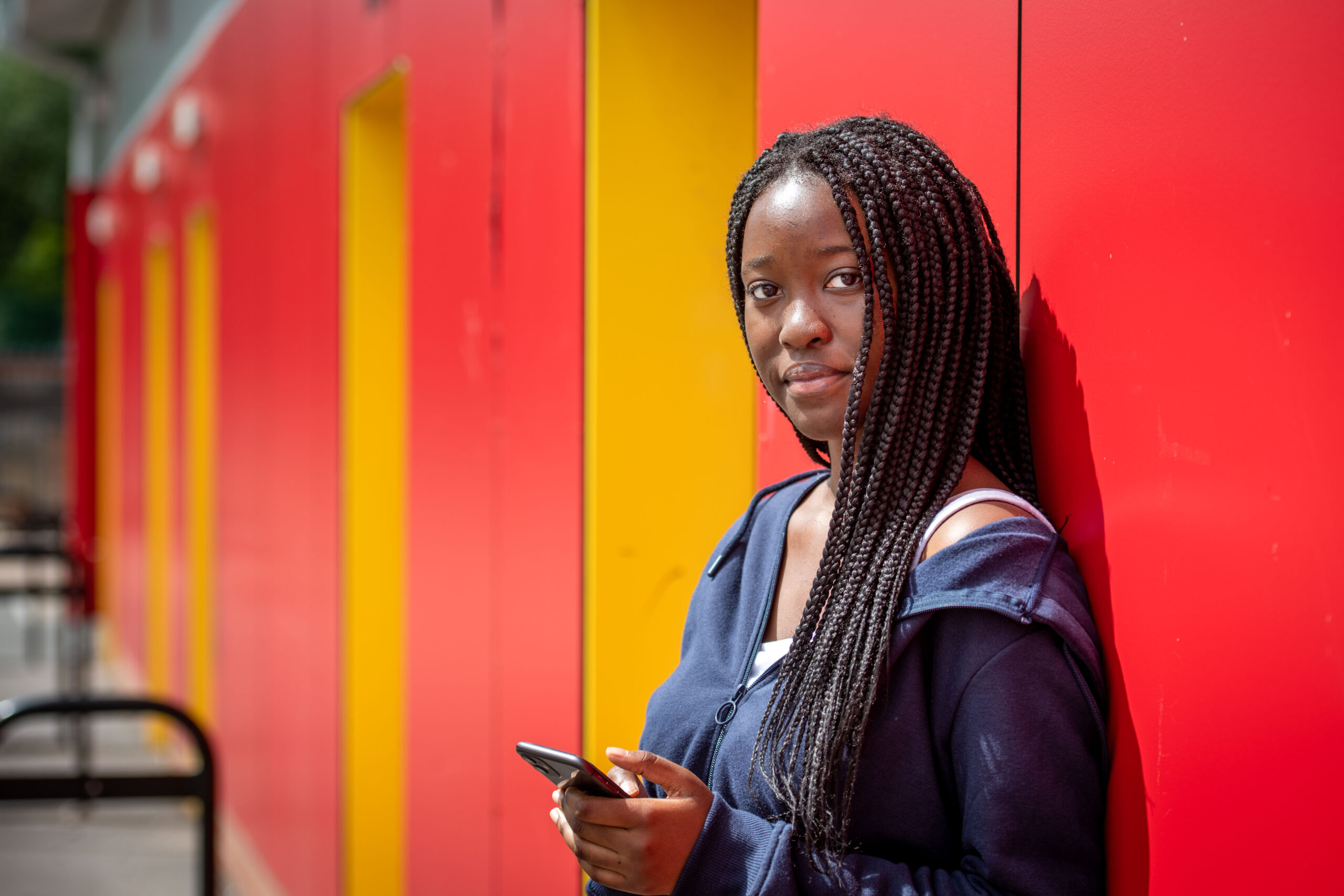 A photograph of Melissa Denga, a college student who is smiles while holding her phone and leaning against a wall.