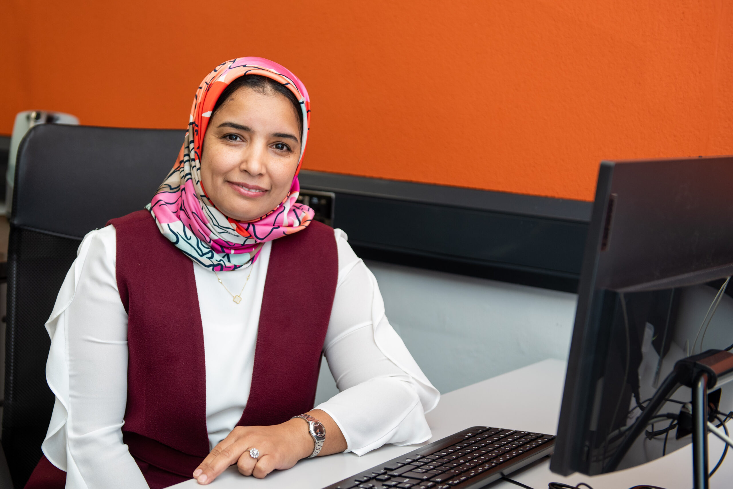 A photograph of Hafida Bouhadi. She is sat at a computer desk with a computer in front of her. She looks at the camera with a smile and rests her left arm on the computer desk.