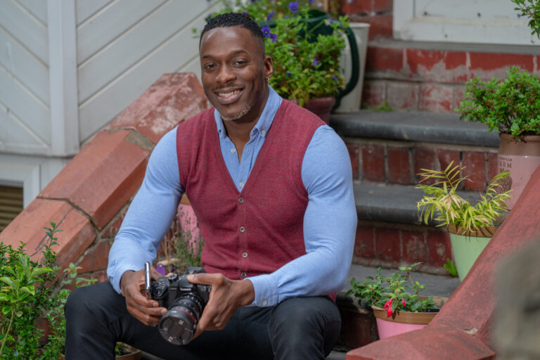 A photograph of David Kemmer-Amoda. He is sat outside on steps while holding a digital camera, and looking at the camera with a smile.