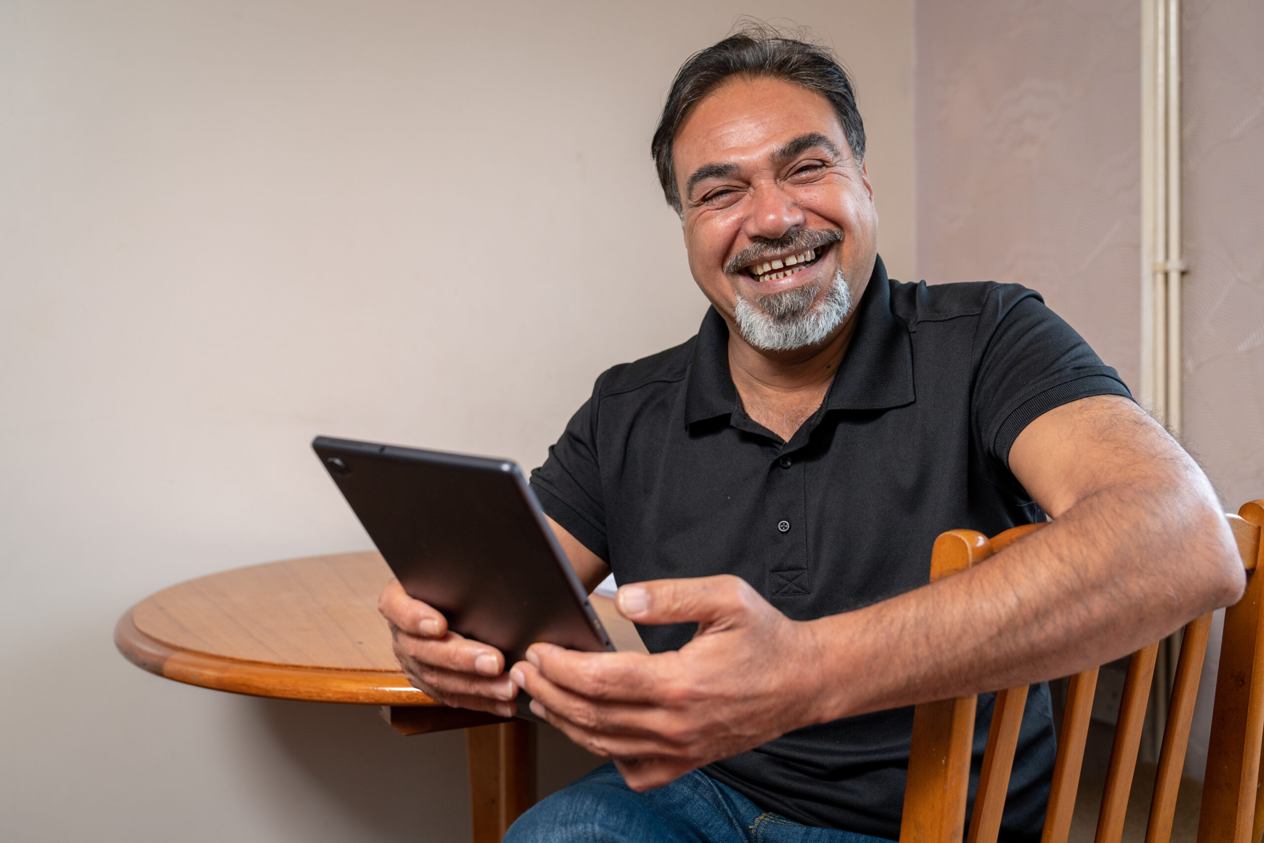 A photograph of Faris Abedalakhwa. He is sat on a chair in front of a circular table. He is smiling while holding a tablet device with both hands in front of him.
