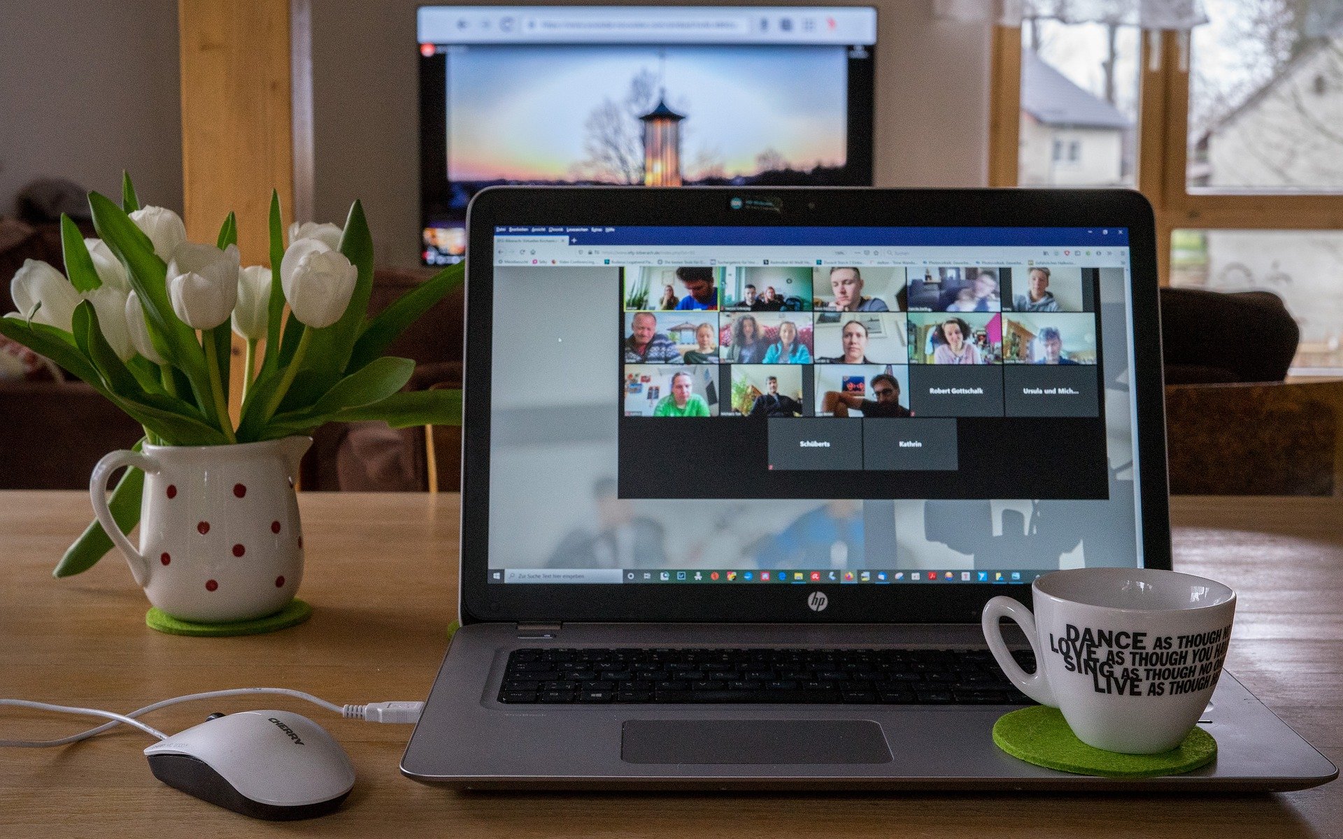 A photograph of a laptop displaying a Zoom meeting on a desk. Also on the desk is a computer mouse, a tea cup and a vase of flowers.