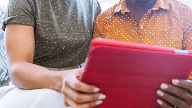 A close up photograph of two people sat down using a tablet. They both hand one hand each on the tablet that is being held.