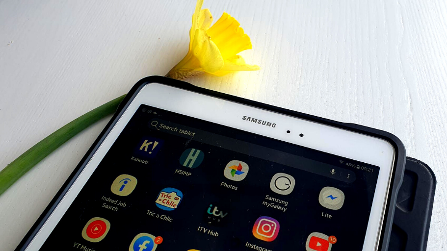 A photograph of a Samsung tablet, laid down on a table, displaying apps on the screen. Next to the tablet is a daffodil.