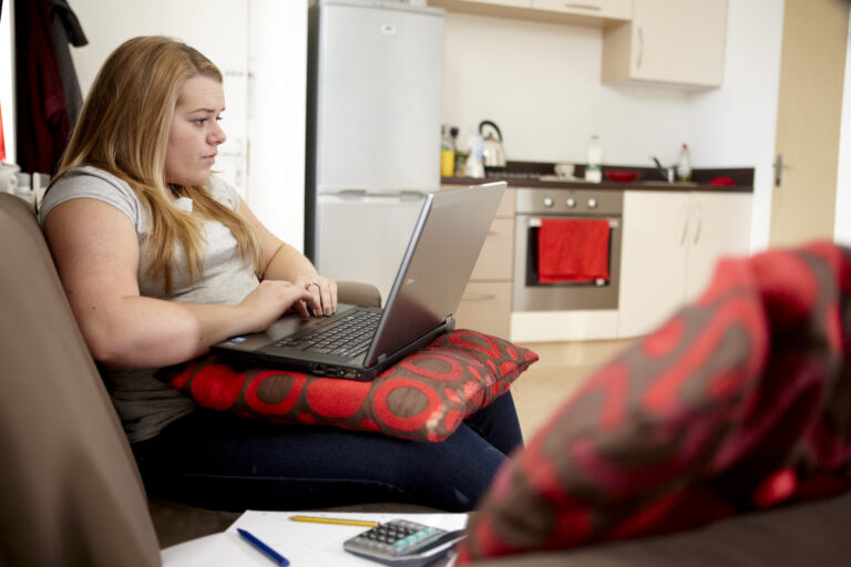 A photograph of a young woman searching for work on laptop. She is sat down on a chair and the laptop is rested on a cushion on her lap.