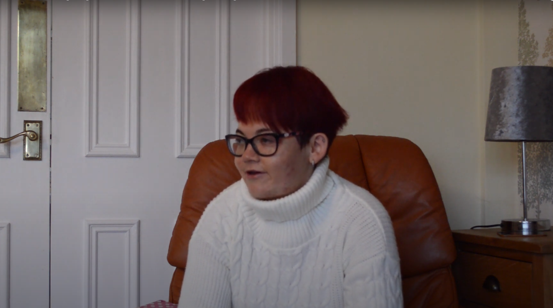 A screenshot from a YouTube video. The image is of Kylie, a device loan scheme beneficiary. She is sat down on a chair in a living room.