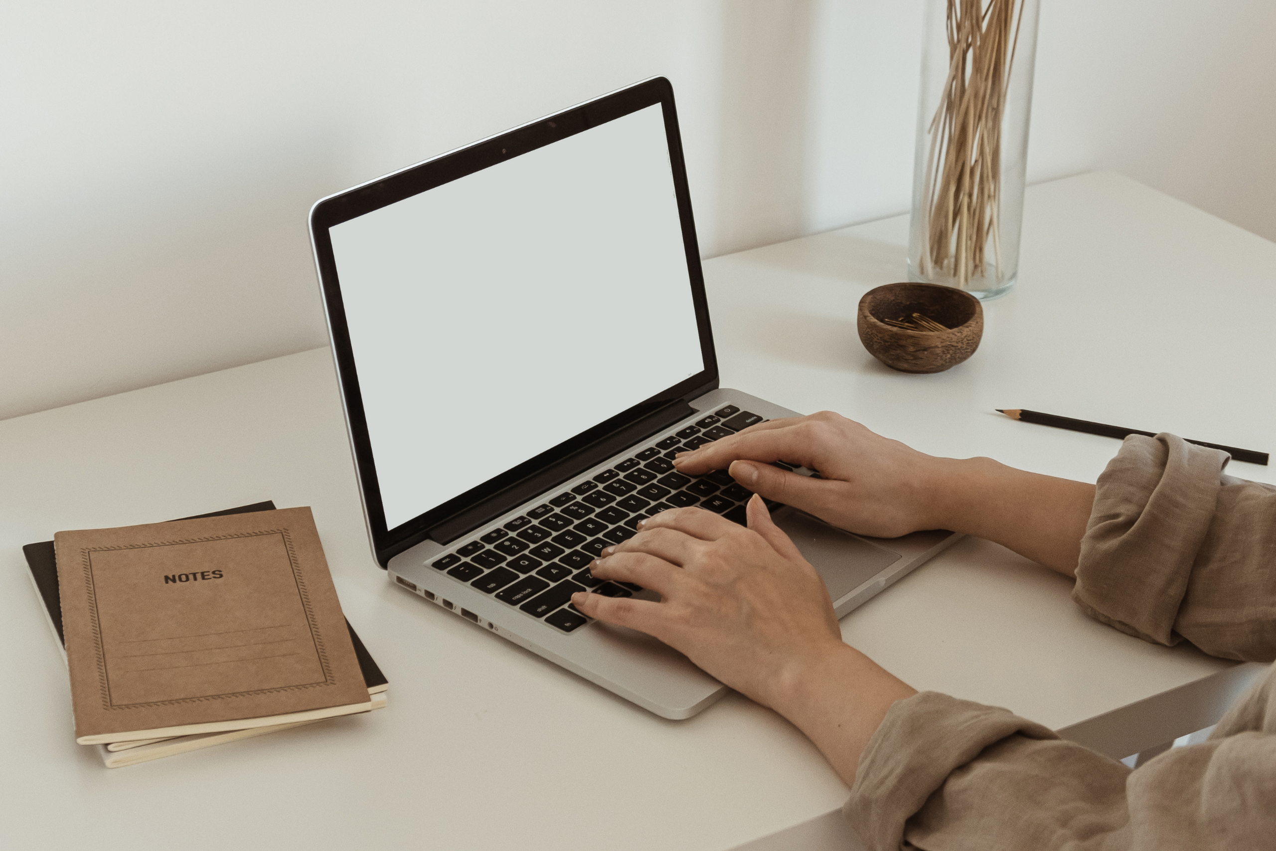 A photograph of two female hands using a laptop. Two stacked notebooks, a pen and some ornaments sit on the desk where the laptop is placed.