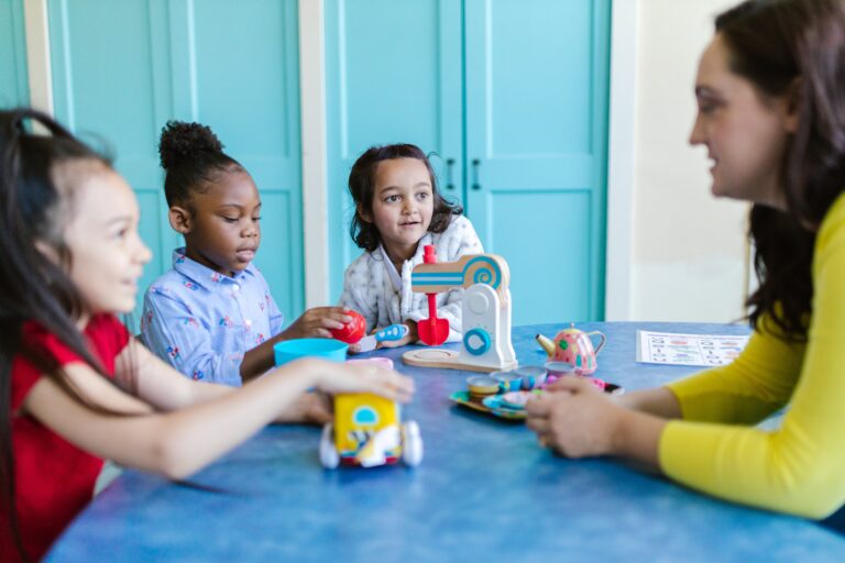 A photograph of a nursery worker talking to three children, all sat around a table. On the table there are children's toys.