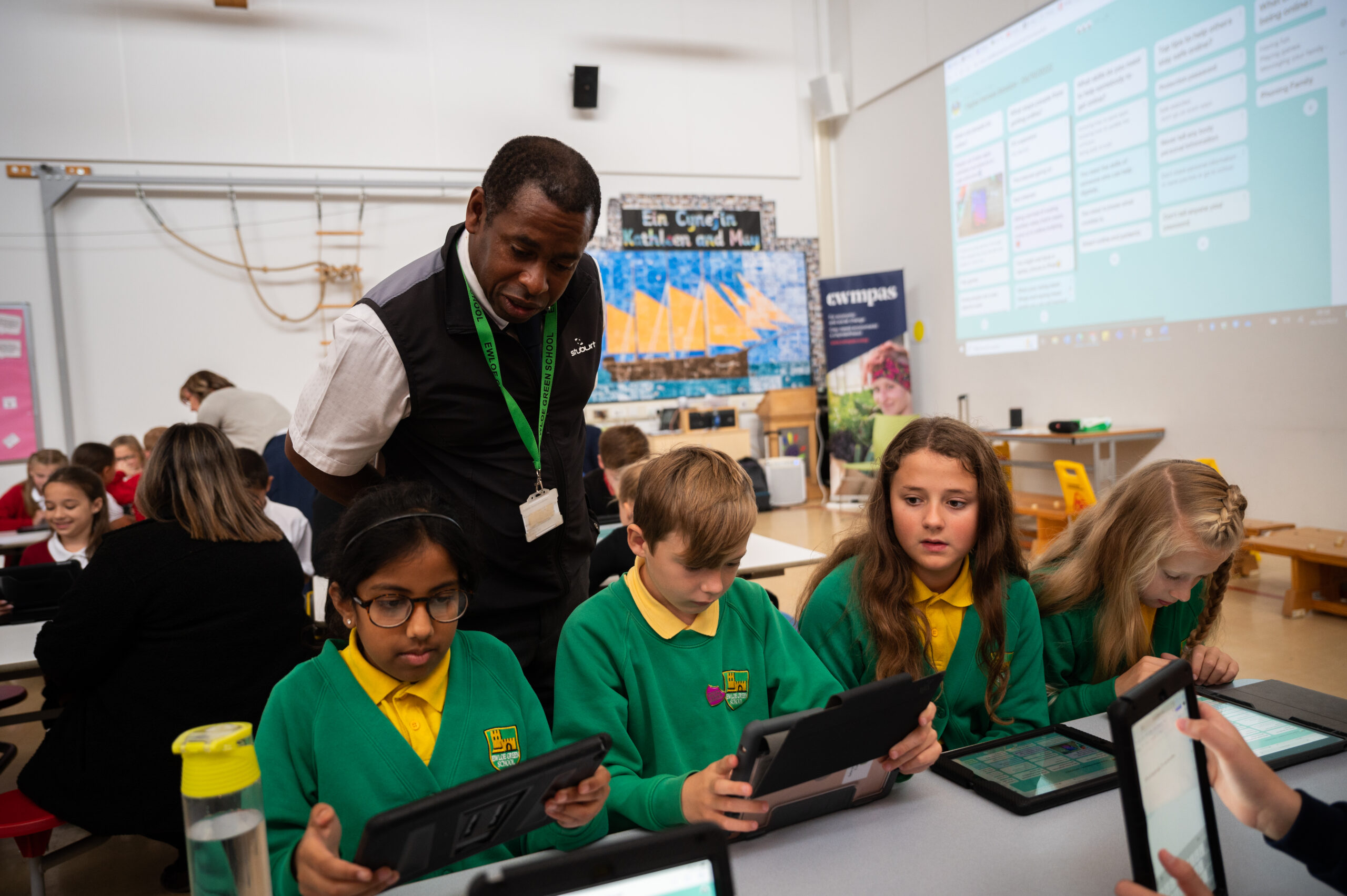 A photograph of a male teacher supervising four Digital Heroes, who are sat at a desk using tablet devices  