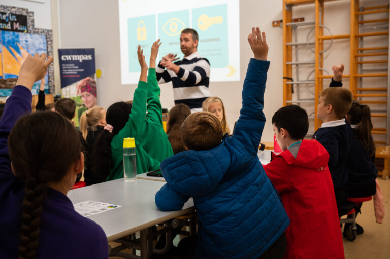 A photograph of a DCW staff member hosting a Digital Heroes training session. Multiple children in the class have their hands raised to ask a question.