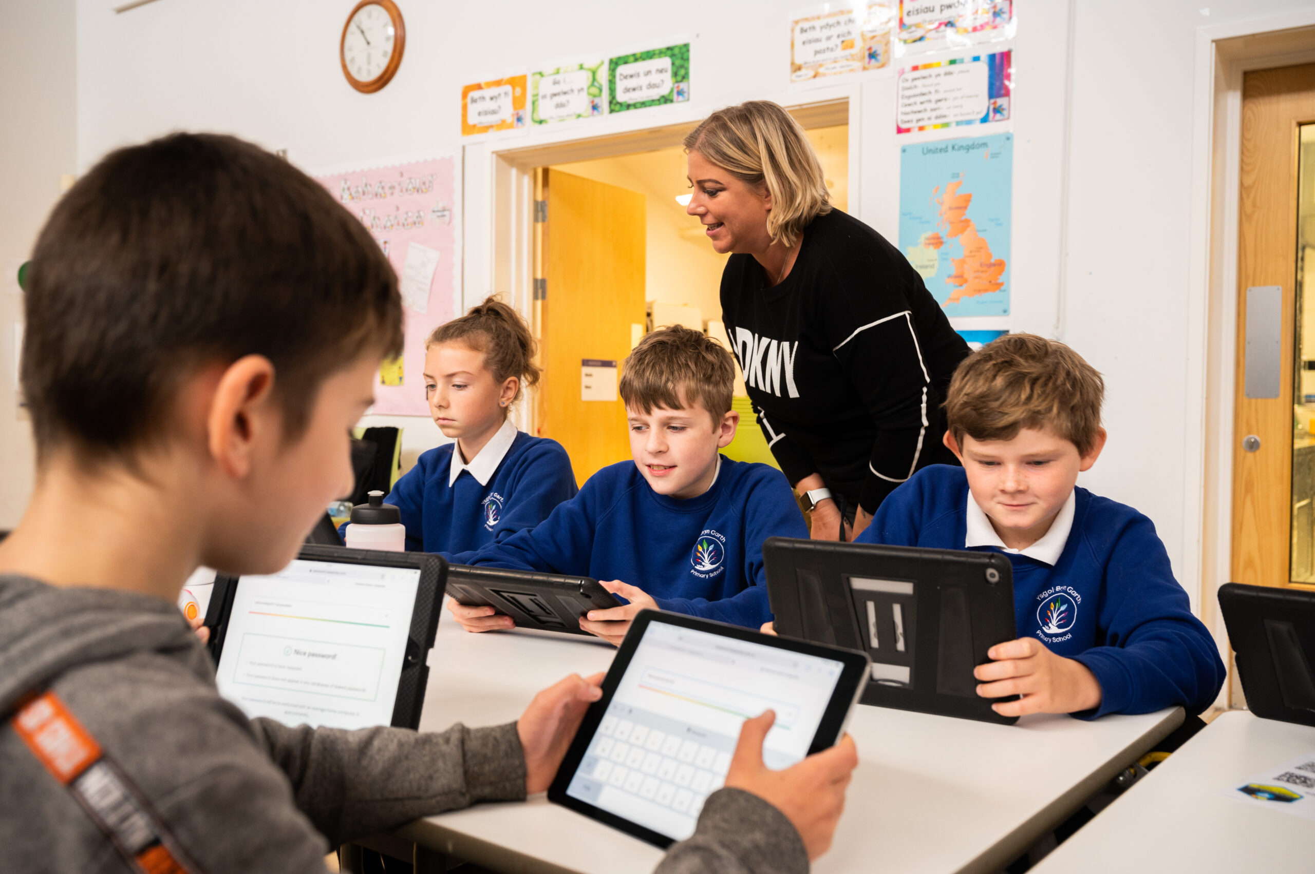 A photograph of four primary school pupils engaging in Digital Heroes training. They're all sat around a desk holding tablet devices while a DCW staff member supervises.