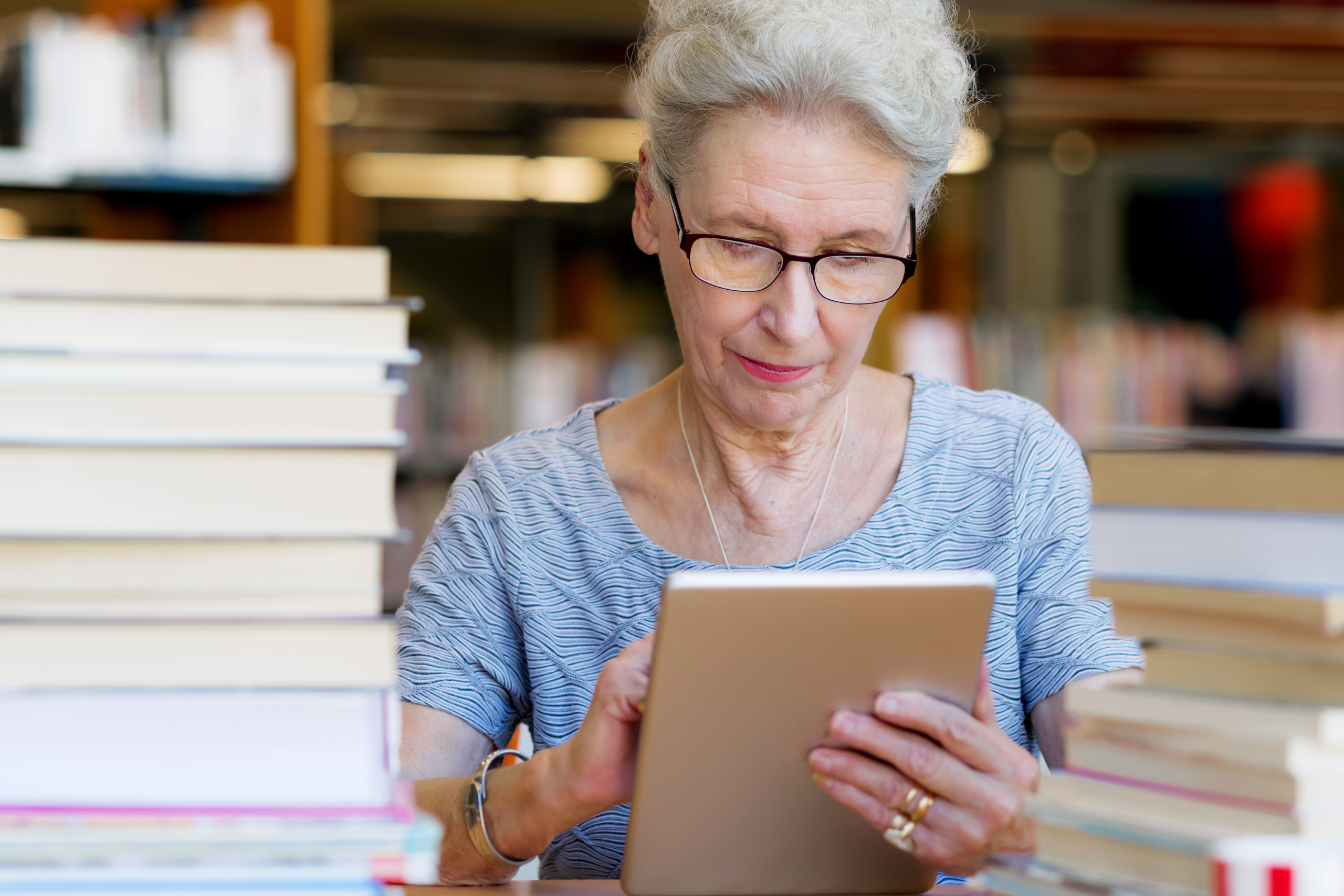 An older woman sits in a library while using a tablet.
