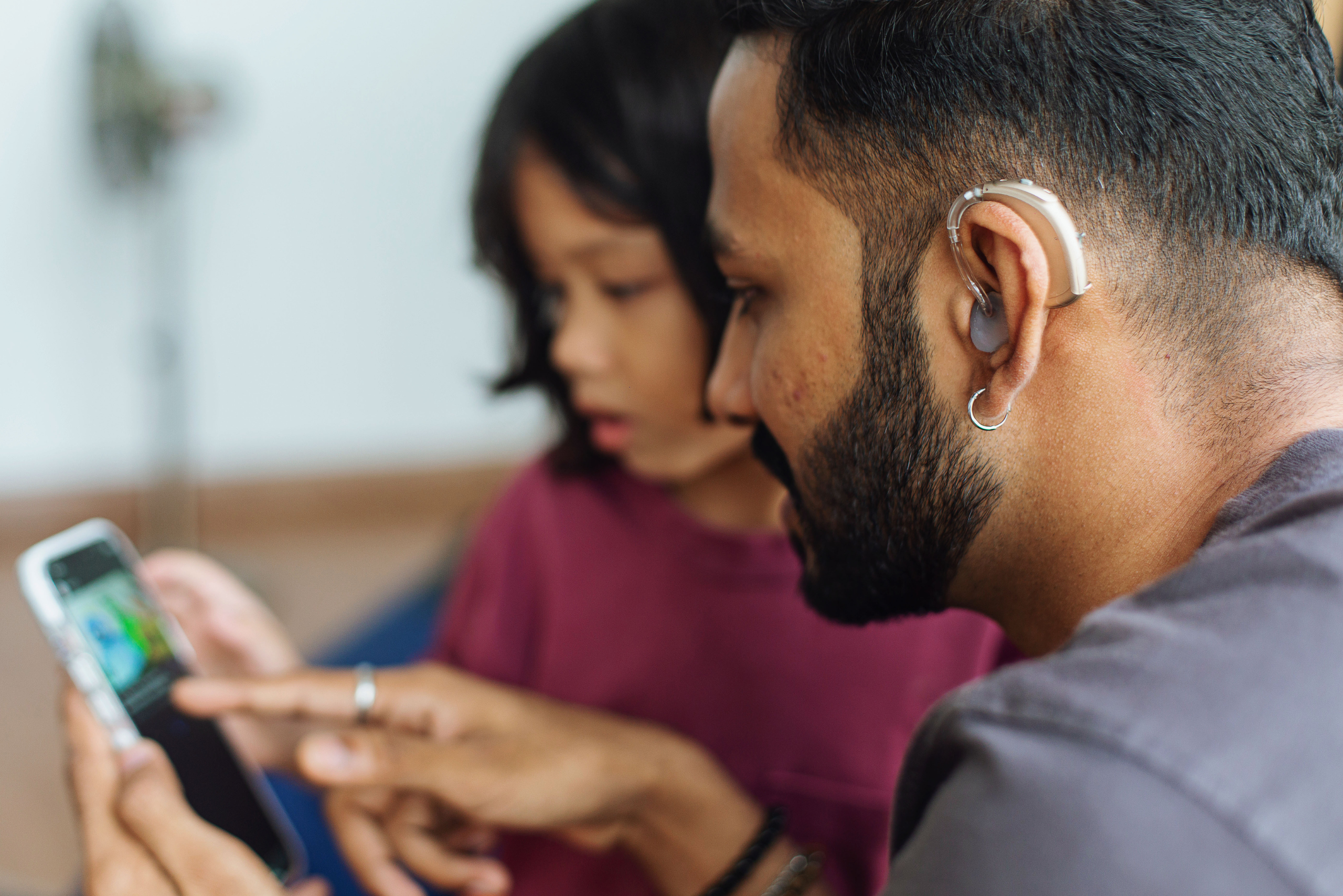 A man using a hearing aid uses a mobile device while a child watches.